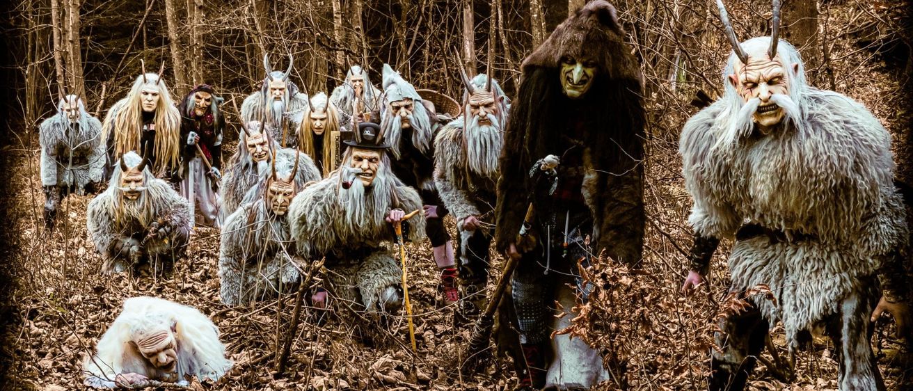 Eine Gruppe von Menschen in Waldkostümen mit Hörnern und Bärten geht durch einen Wald. Einige knien, andere stehen und alle schauen nach vorne.