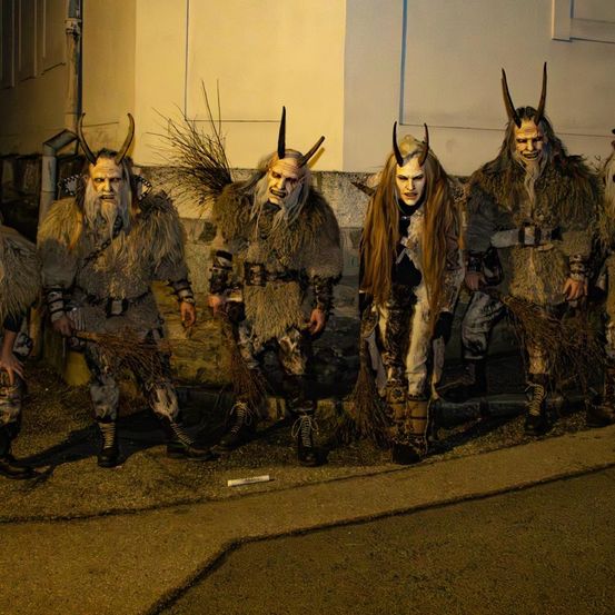 Fünf Personen in Hörnerkostümen und Masken stehen in einer Reihe auf einem Bürgersteig bei Nacht. Sie halten Besen. Das Gebäude hinter ihnen hat eine weiße Wand und eine Rohrleitung.