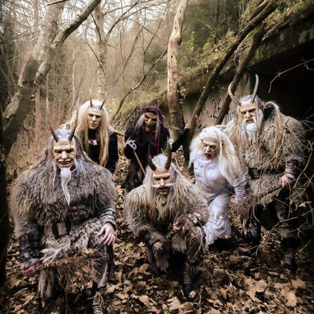 Eine Gruppe von Menschen in Hörnermasken posiert in einem Wald mit einer Betonmauer im Hintergrund. Sie halten Stöcke mit Blättern.