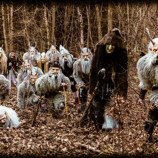 Eine Gruppe von Menschen in Kostümen steht in einem Wald. Sie haben lange Bärte und Hörner. Sie scheinen eine Zeremonie durchzuführen.