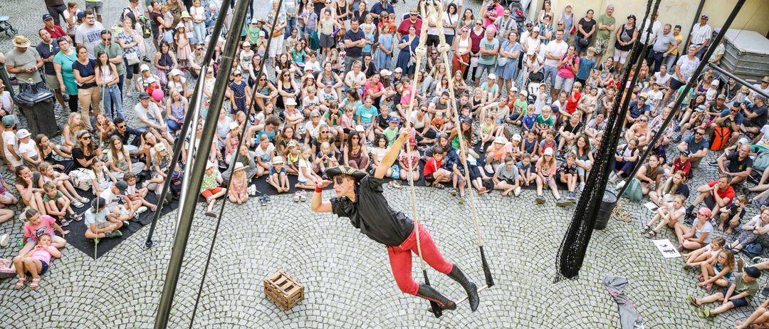 A performer in a black and red outfit is hanging from ropes in the middle of a crowd, possibly performing a circus act.