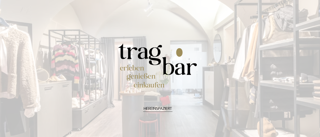 Eine Innenwerbung für 'trag bar' lädt zum Erleben, Genießen und Einkaufen ein. Die Umgebung zeigt ein modernes Interieur mit Holzboden, rundem Tisch und weißer Wand mit Spiegel.