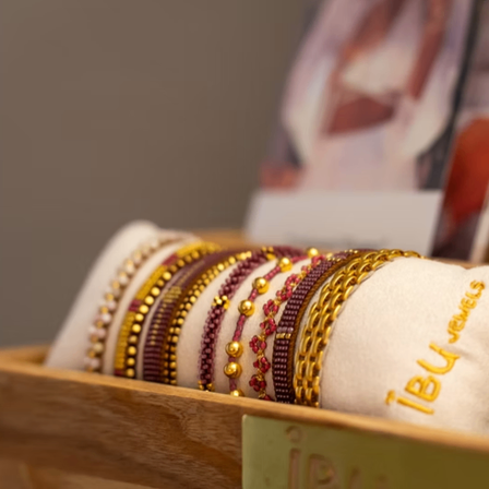 Eine Holzkiste zeigt mehrere Armbänder mit Perlen, mit einem Etikett, das '18U Jewels' liest. Der Hintergrund ist verschwommen und zeigt eine Zeitschrift und eine Person.