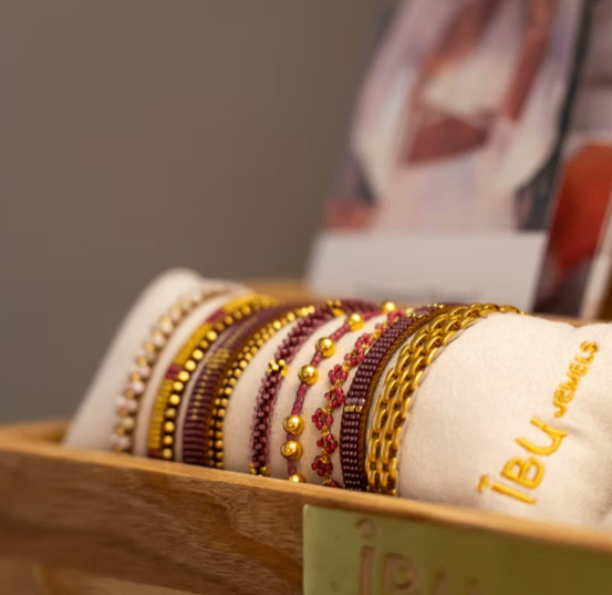 Eine Holzkiste zeigt mehrere Armbänder mit Perlen, mit einem Etikett, das '18U Jewels' liest. Der Hintergrund ist verschwommen und zeigt eine Zeitschrift und eine Person.