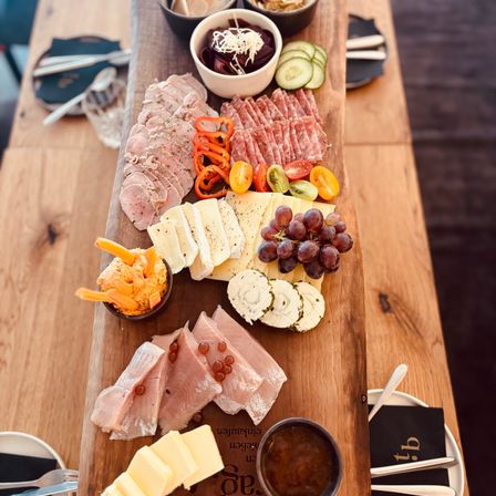 Ein Holzbrett zeigt verschiedene Fleischsorten, Käse und Gemüse. In der Nähe befinden sich Schalen mit Salat und Hummus.
