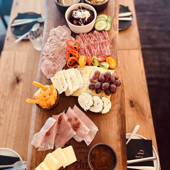 Ein Holzbrett zeigt verschiedene Fleischsorten, Käse und Gemüse. In der Nähe befinden sich Schalen mit Salat und Hummus.