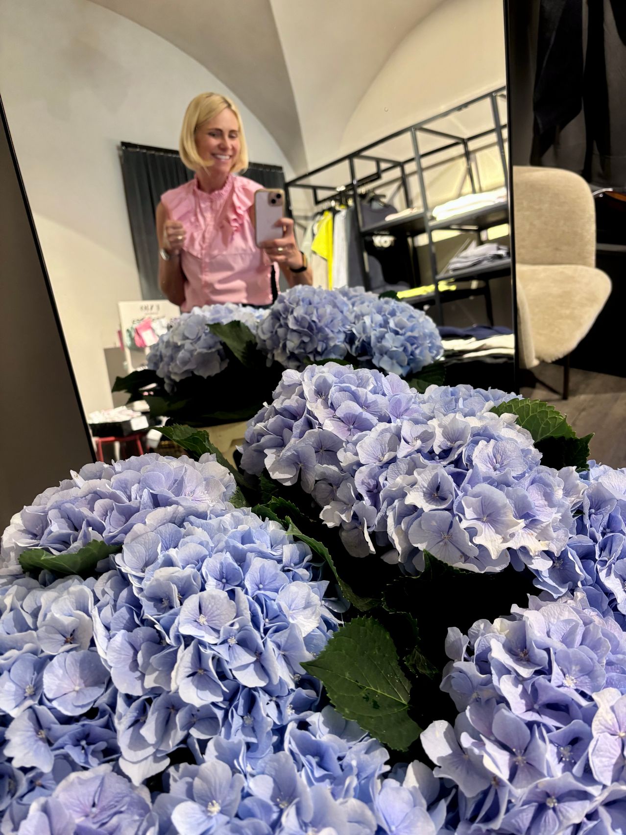 Eine Frau macht ein Selfie vor einem Spiegel mit einem Strauß blauer Hortensien im Vordergrund.