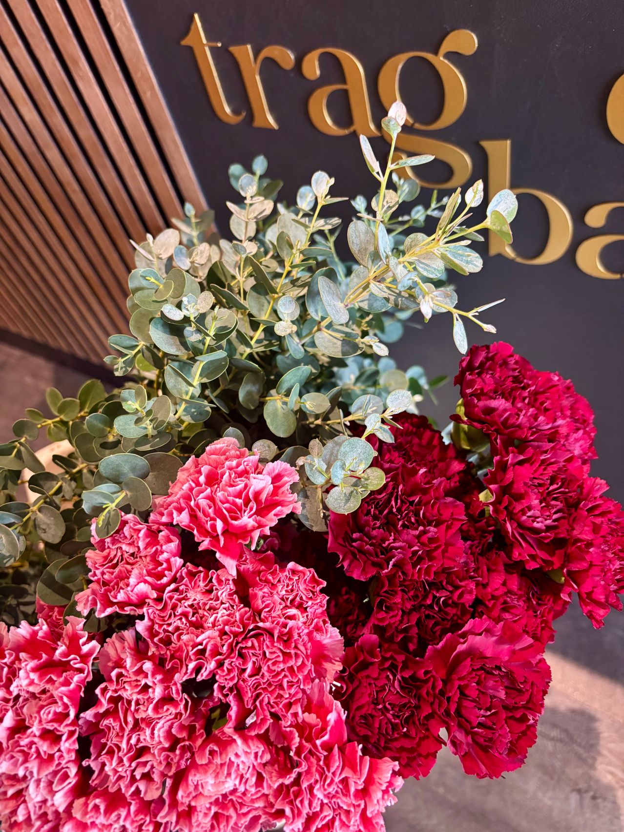 Ein Nahaufnahme einer farbenfrohen Blumenarrangement mit rosa Nelken und grünen Blättern, vor einer schwarzen Wand mit goldenem Text dargestellt.