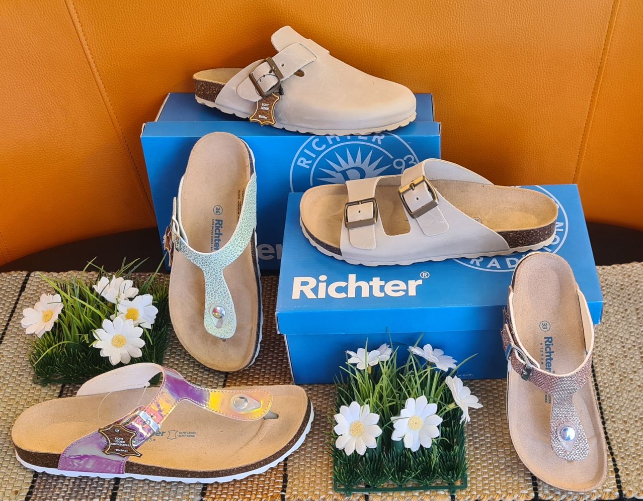 Mehrere Paare Richter-Sandalen in verschiedenen Farben sind auf einem Tisch ausgestellt. Die Sandalen liegen auf blauen Schachteln, mit je zwei Blumen auf jeder Seite des Tisches.