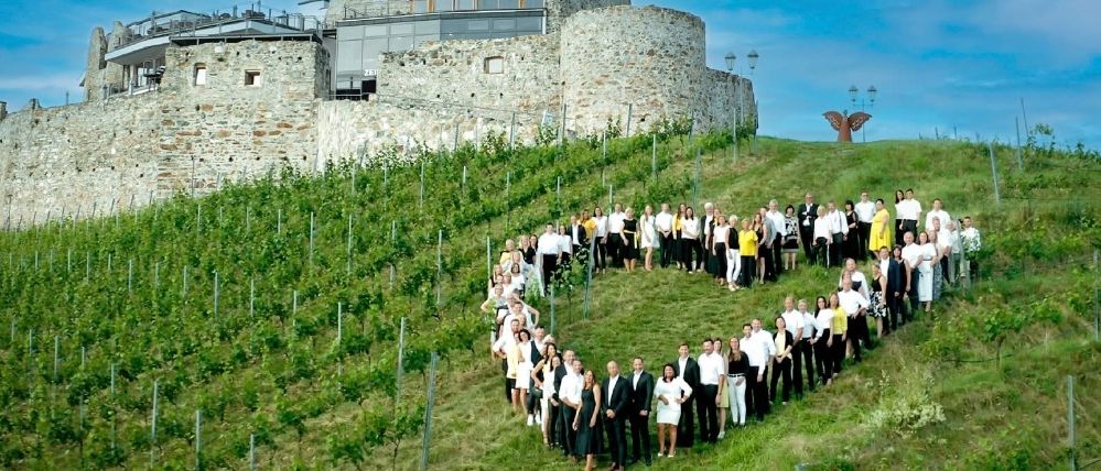 Eine Gruppe formell gekleideter Menschen bildet eine Herzform auf einem grünen Hügel, mit einer Burg im Hintergrund.