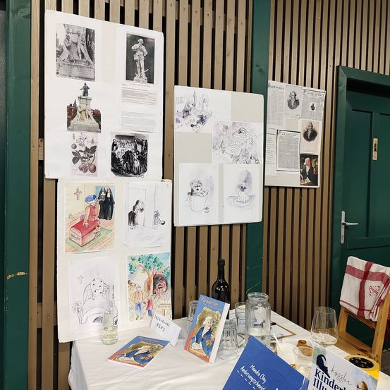 Ein Tisch zeigt Bücher und Weinflaschen, wobei eine Holzwand mit verschiedenen Zeichnungen, Papieren und Fotos geschmückt ist, und eine geschlossene grüne Tür auf der rechten Seite.