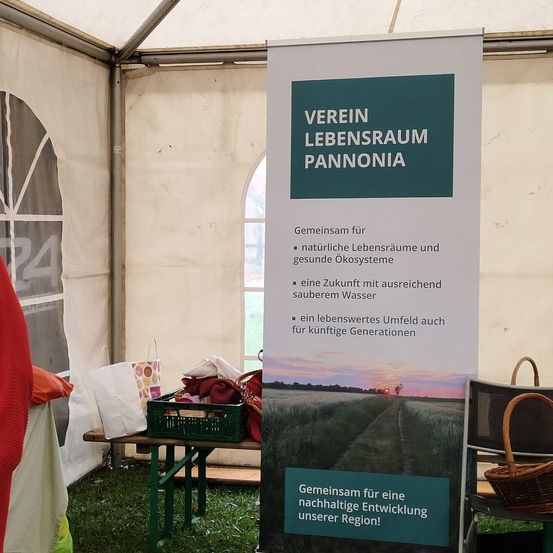 Ein Plakat für den Verein Lebensraum Pannonia steht in einem Zelt und fördert natürliche Lebensräume und sauberes Wasser für zukünftige Generationen. Das Plakat zeigt ein ländliches Landschaftsbild.