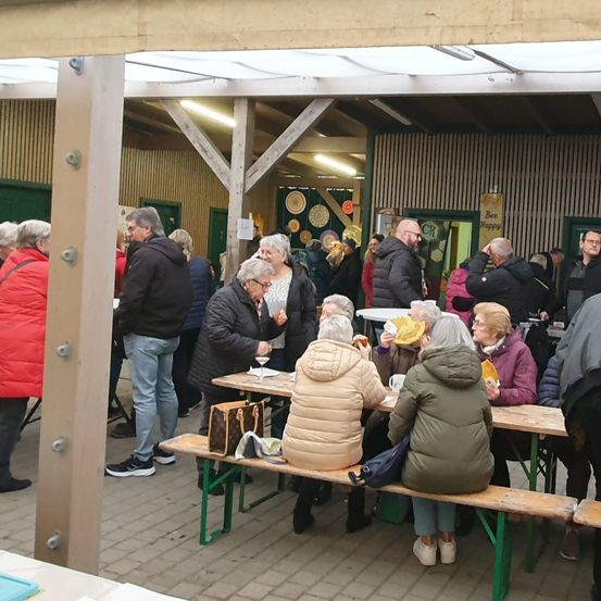 Eine Gruppe von Menschen versammelt sich in einem Außenbereich. Einige sitzen auf Bänken, andere stehen. Die Tische sind mit Essen und Getränken gedeckt. Der Bereich befindet sich unter einem Dach mit Holzbalken.