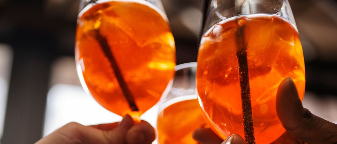 Drei Hände halten Weingläser mit orangefarbenen Getränken und prosten gemeinsam, mit einem unscharfen Hintergrund.