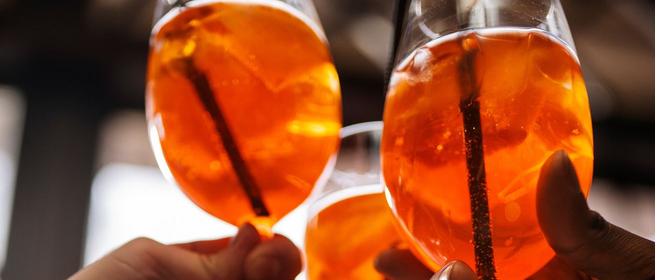 Drei Hände halten Weingläser mit orangefarbenen Getränken und prosten gemeinsam, mit einem unscharfen Hintergrund.