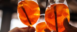 Drei Hände halten Weingläser mit orangefarbenen Getränken und prosten gemeinsam, mit einem unscharfen Hintergrund.
