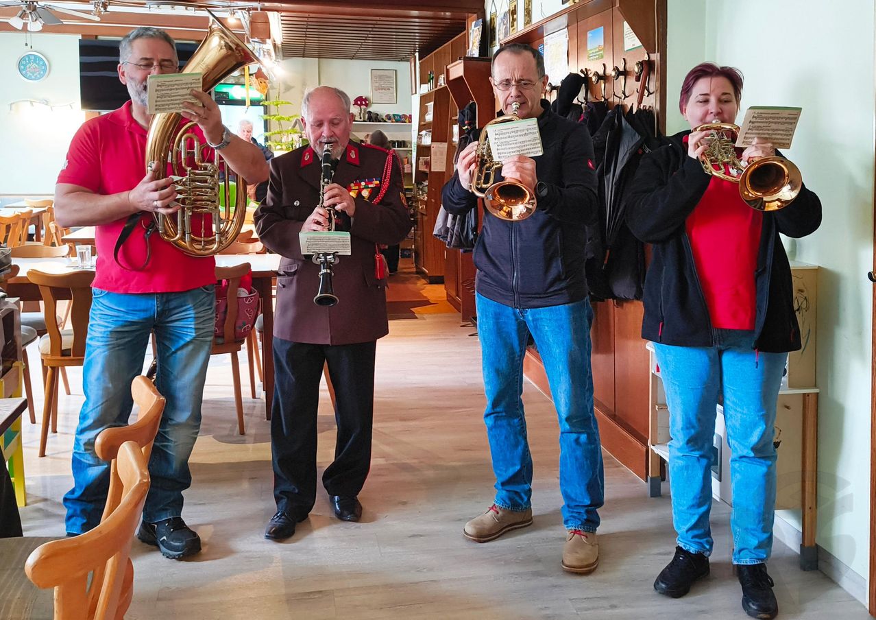 Vier Personen, zwei Männer und zwei Frauen, stehen in einem Raum und spielen Blechblasinstrumente. Sie sind alle lässig gekleidet. Hinter ihnen befinden sich Holzregale mit verschiedenen Gegenständen.