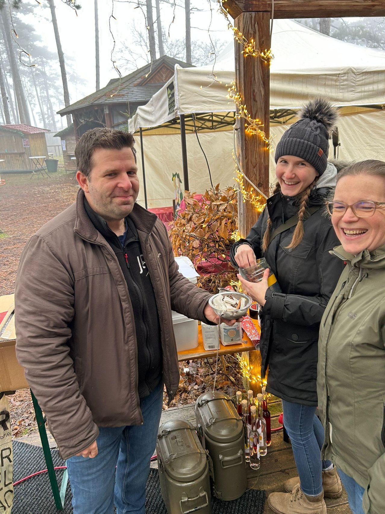 Drei Personen, zwei Frauen und ein Mann, stehen in einem nebligen Außenbereich, möglicherweise auf einem Markt. Der Mann hält eine Schüssel und reicht sie einer der Frauen. Sie alle lächeln. Hinter ihnen steht ein Zelt mit einem Holzpfahl, und mehrere Flaschen stehen auf einem Tisch.