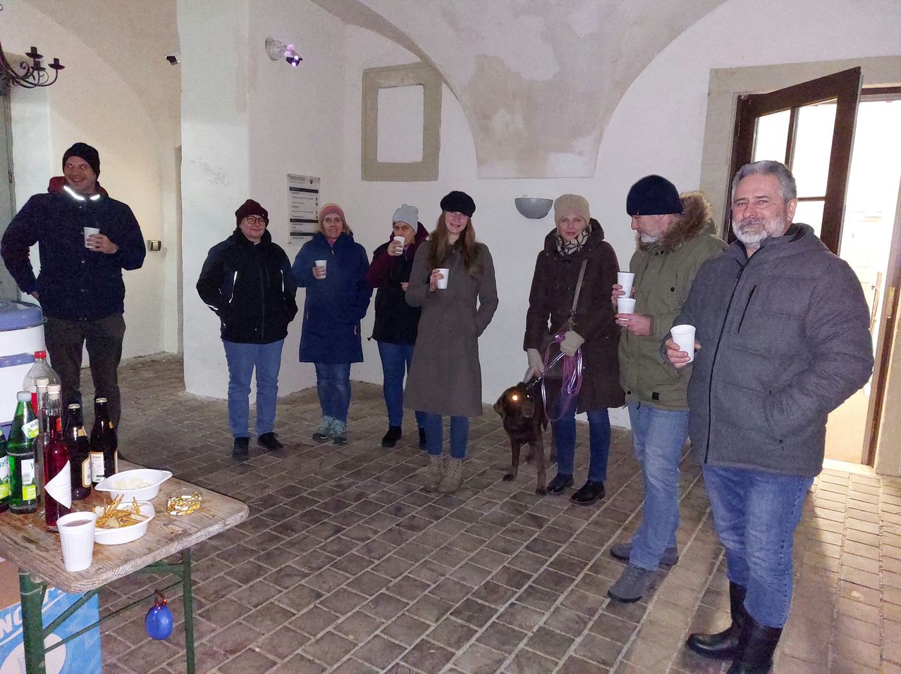 Eine Gruppe von Menschen steht in einem Raum mit Ziegelböden, in Winterkleidung. Sie halten Tassen und lächeln. Ein Hund steht neben einer Frau. Ein Tisch steht vor ihnen.