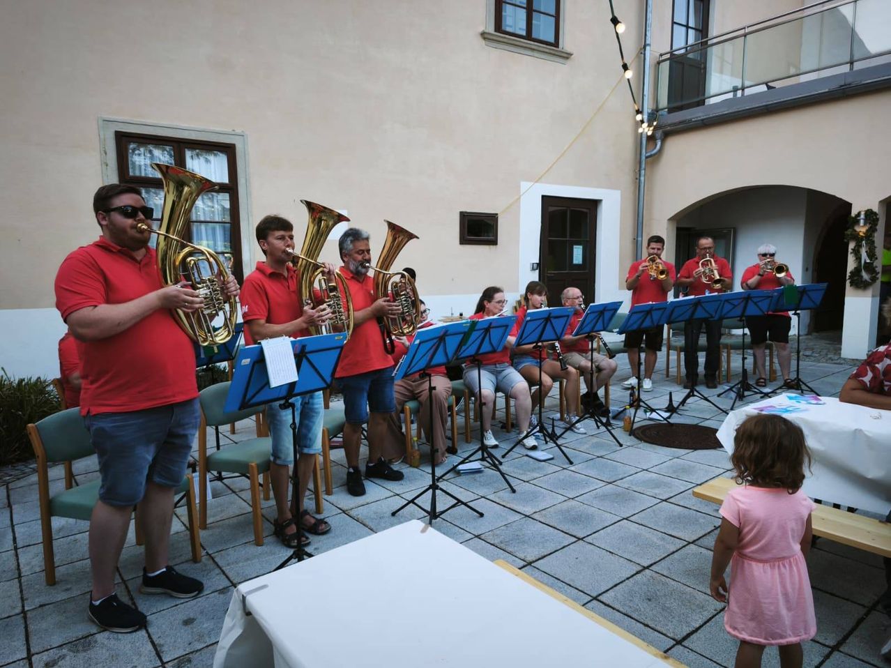 Eine Gruppe von Musikern in roten Hemden und Shorts spielt Blechblasinstrumente in einer Außenumgebung. Sie stehen vor einem Gebäude mit Balkon und Lichterketten.