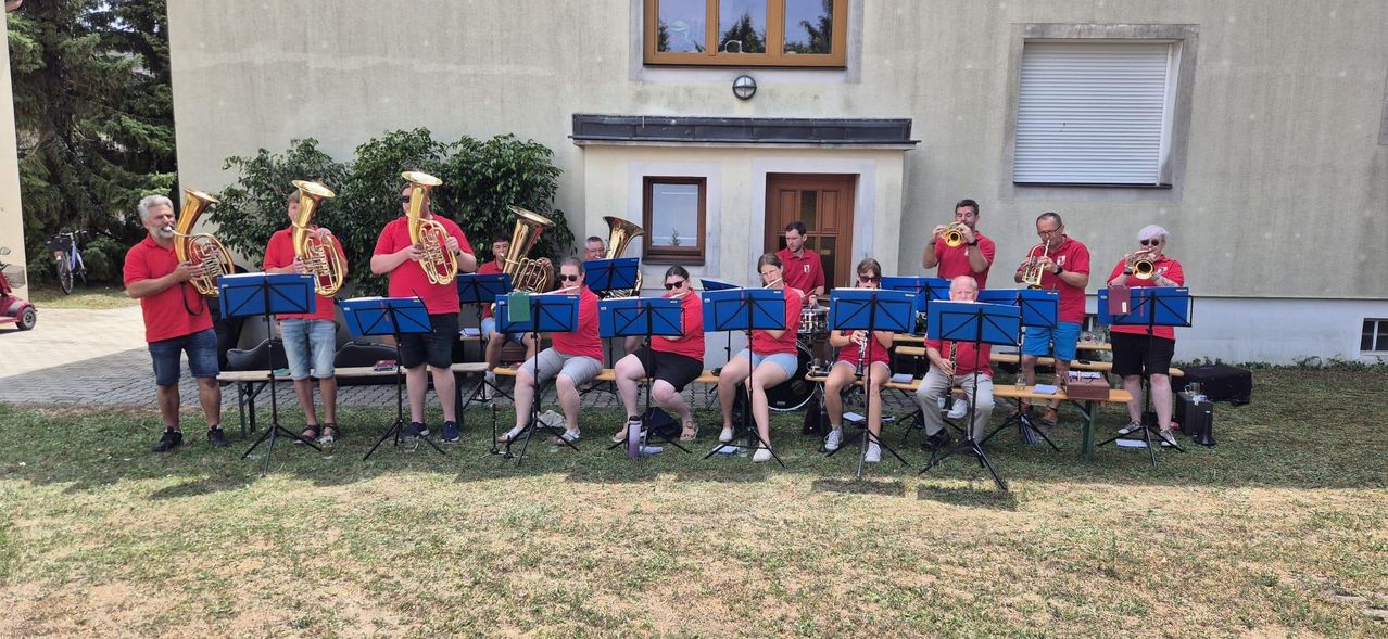 Eine Blechbläsergruppe sitzt auf einer Bank vor einem Gebäude, wobei die Musiker verschiedene Instrumente wie Trompeten, Tuben und Klarinetten spielen.