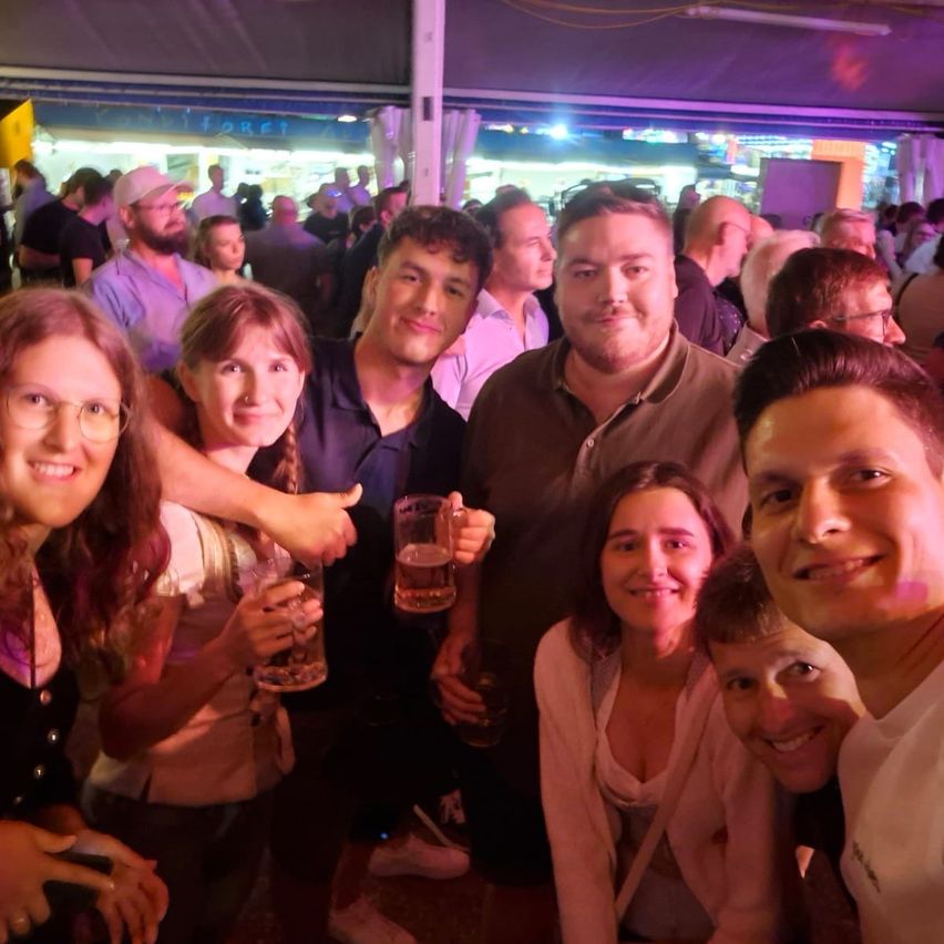 A group of people are smiling and posing for a photo at a crowded event, holding glasses of beer.