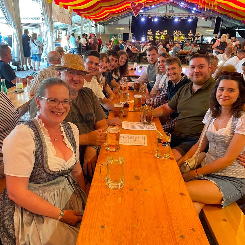 A group of people sitting at a long table, smiling and posing for a photo. They are holding glasses of beer. Behind them, there are more people sitting and standing, and a stage with a band.