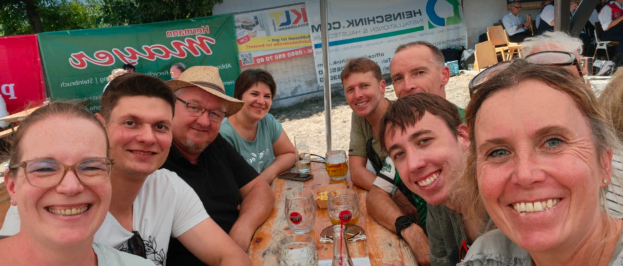 A group of people are sitting around a table with glasses of beer, smiling for a photo. They are under a tent with banners in the background.