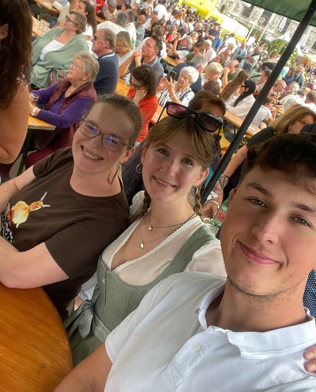 Three people smiling for a photo at an outdoor event, with a crowd of people sitting at tables in the background.