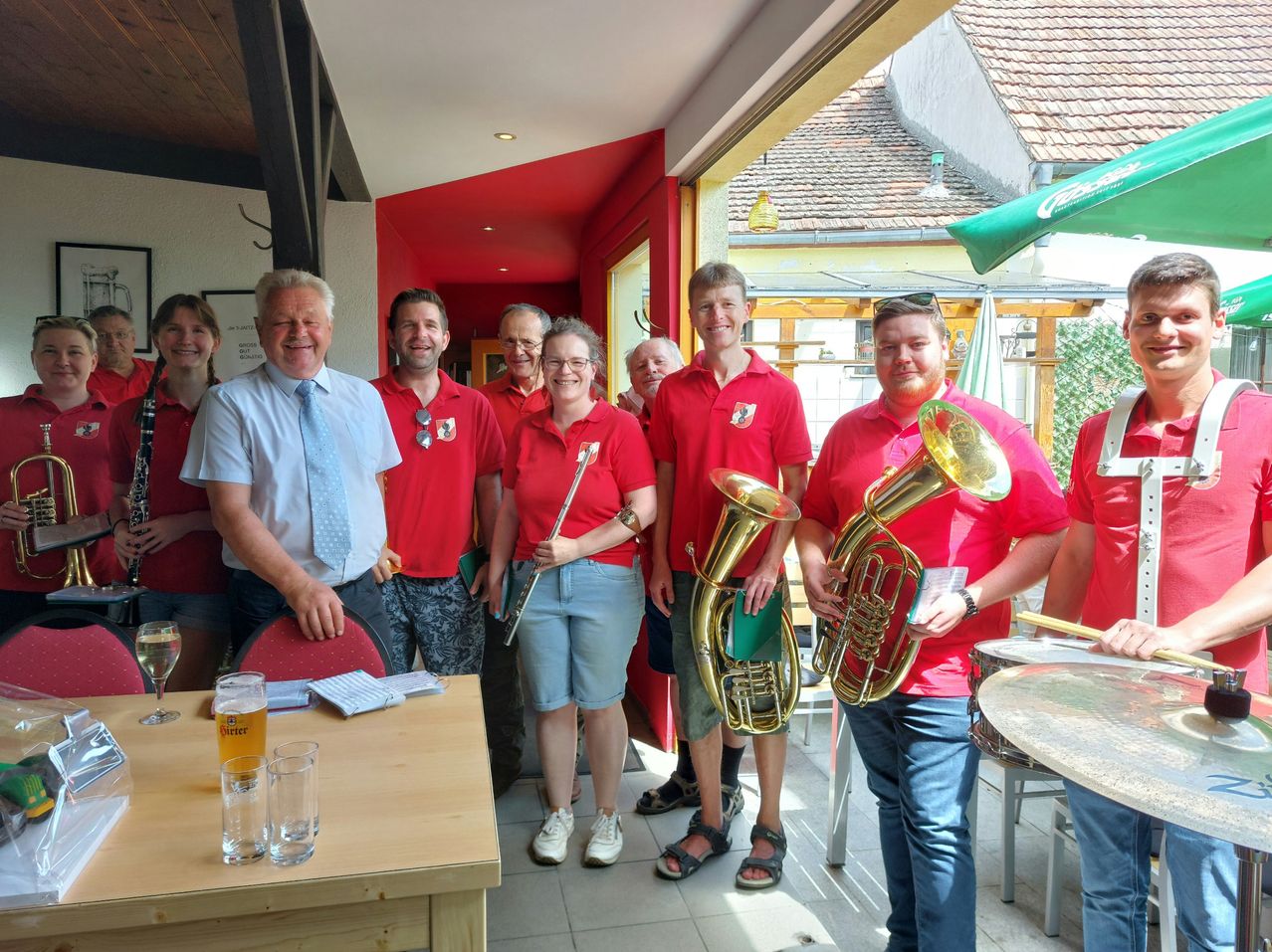 Eine Gruppe von Menschen, einige in roten Hemden, posiert mit Musikinstrumenten in einem Restaurant. Ein Tisch mit Gläsern und Papieren ist in der Nähe.