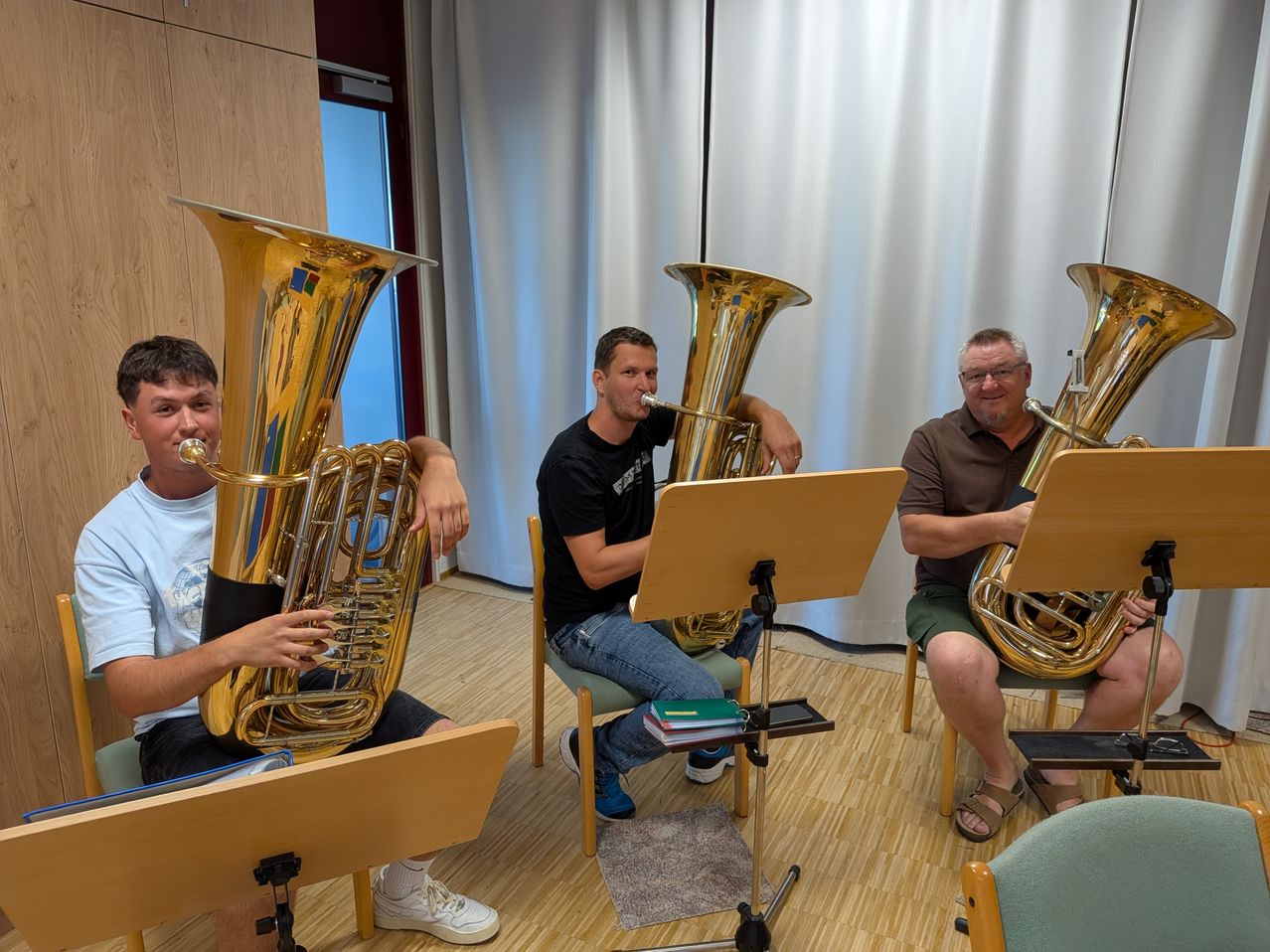 Three men are seated in chairs, playing large brass tubas. The man on the left wears a white shirt and shorts, the middle man a black shirt and jeans, and the man on the right a brown shirt and green shorts.