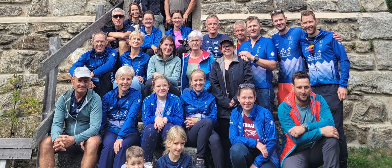 Eine Gruppe von Menschen in identischen blauen Jacken mit Logos, die vor einem steinernen Gebäude sitzen und stehen. Einige tragen Hüte und Sonnenbrillen. Hinter ihnen stehen zwei Personen in der Nähe des Eingangs.