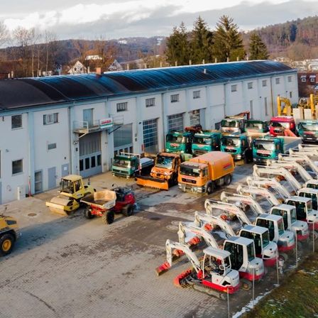 Ein großes weißes Gebäude mit einem blauen Dach beherbergt eine Flotte von Baufahrzeugen, darunter Bagger, Lastwagen und Bulldozer, die vor ihm in einer organisierten Weise geparkt sind.
