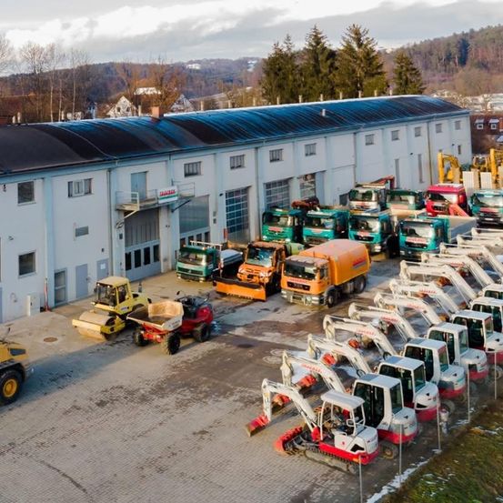 Ein großes weißes Gebäude mit einem blauen Dach beherbergt eine Flotte von Baufahrzeugen, darunter Bagger, Lastwagen und Bulldozer, die vor ihm in einer organisierten Weise geparkt sind.