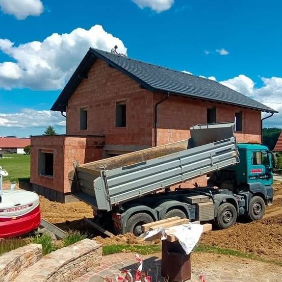 Eine Baustelle mit einem großen Backsteinhaus im Bau, einem Kipplaster, der Erde entlädt, und einer Person auf dem Dach.