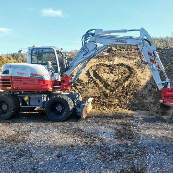 Ein roter und weißer Bagger mit der Modellnummer TB295W steht auf einer Kiesoberfläche und gräbt in einen Haufen Erde.