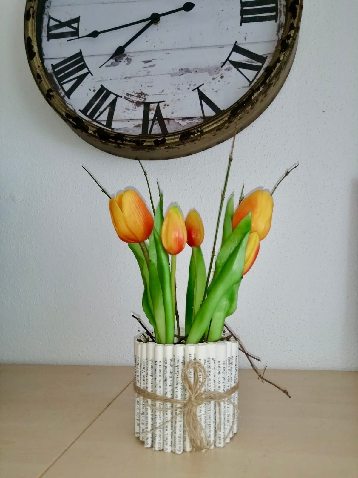 Ein aus Zeitungen gerollter Blumenstrauß hält mehrere gelbe Tulpen auf einem Tisch mit einer weißen Wand und einer alten Uhr im Hintergrund.