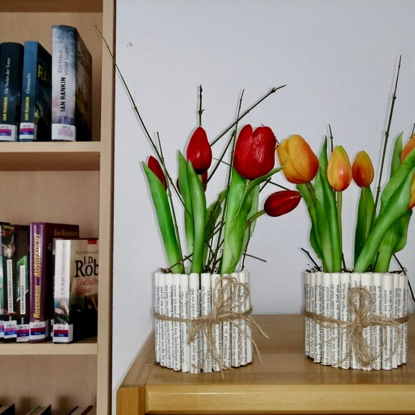 Zwei Vasen mit bunten Tulpen stehen auf einem holzfarbenen Tisch. Die Vasen bestehen aus gerollten Zeitungen. Hinter dem Tisch ist eine weiße Wand und ein mit Büchern gefüllter Bücherregal zu sehen.