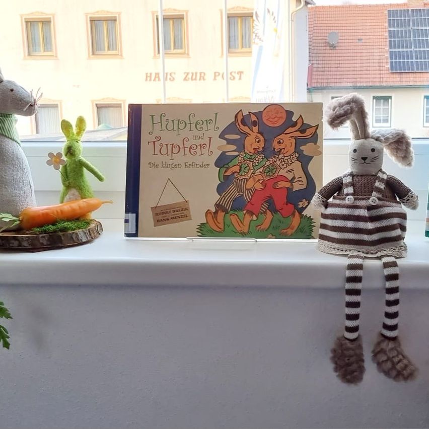 Auf einem Fenstersims steht ein Buch mit dem Titel 'Hupferl und Tupferl' zusammen mit zwei Plüschhasen. Eine Karotte liegt auf einem Baumstumpf, und ein grünes Hasenspielzeug steht in der Nähe.