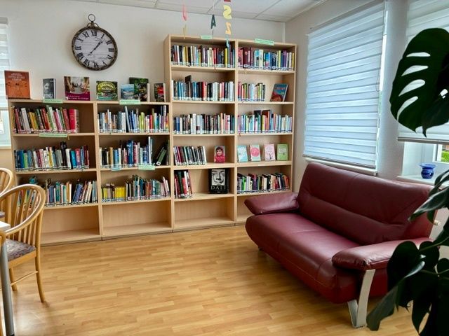 Eine gemütliche Leseecke mit Holzboden, einem roten Ledersofa und einem mit Büchern gefüllten Bücherregal. Eine Uhr und Pflanzen schmücken den Raum.