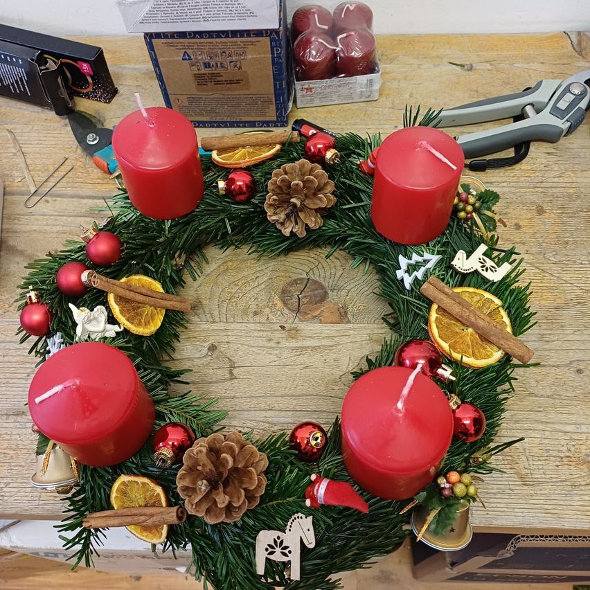 Ein Kranz mit roten Kerzen, Tannenzapfen und Ornamenten auf einer Holzoberfläche. Eine Glocke, Scheren und eine Schachtel sind in der Nähe.