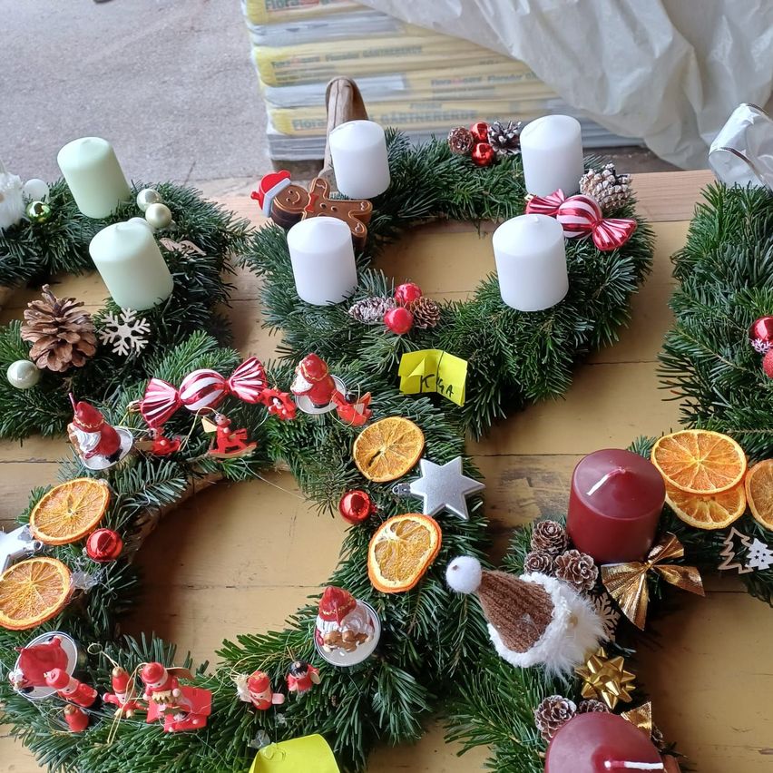 Vier Weihnachtskränze sind auf einem Holztisch angeordnet. Sie haben grüne Zweige, Kerzen, Orangen, Sterne und Weihnachtsmannfiguren. Einige haben Bänder und Tannenzapfen. Ein gelber Zettel ist an einem Kranz befestigt.