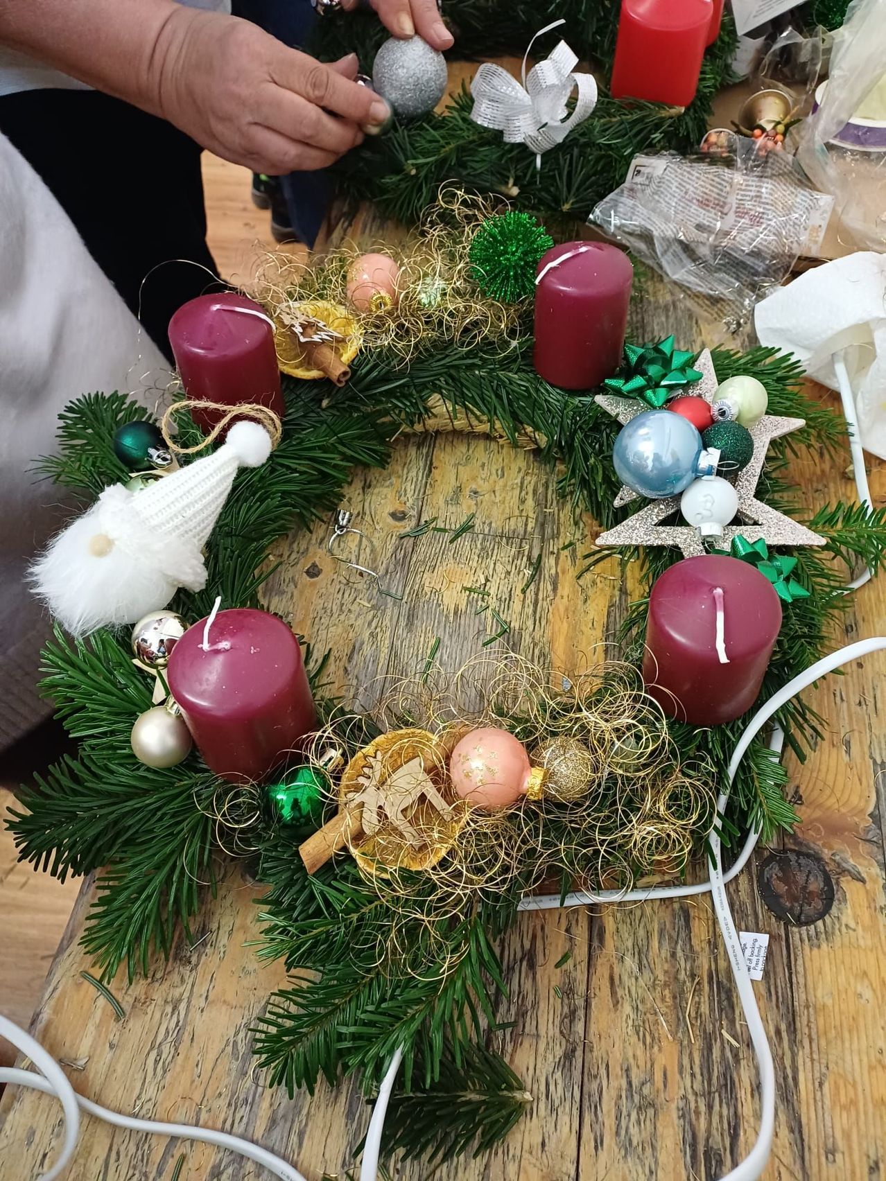 Ein Holztisch mit einem aus Kiefernnadeln gefertigten Kranz, geschmückt mit roten Kerzen, silbernem Tinsel und verschiedenen Ornamenten wie Sternen und Tannenzapfen.