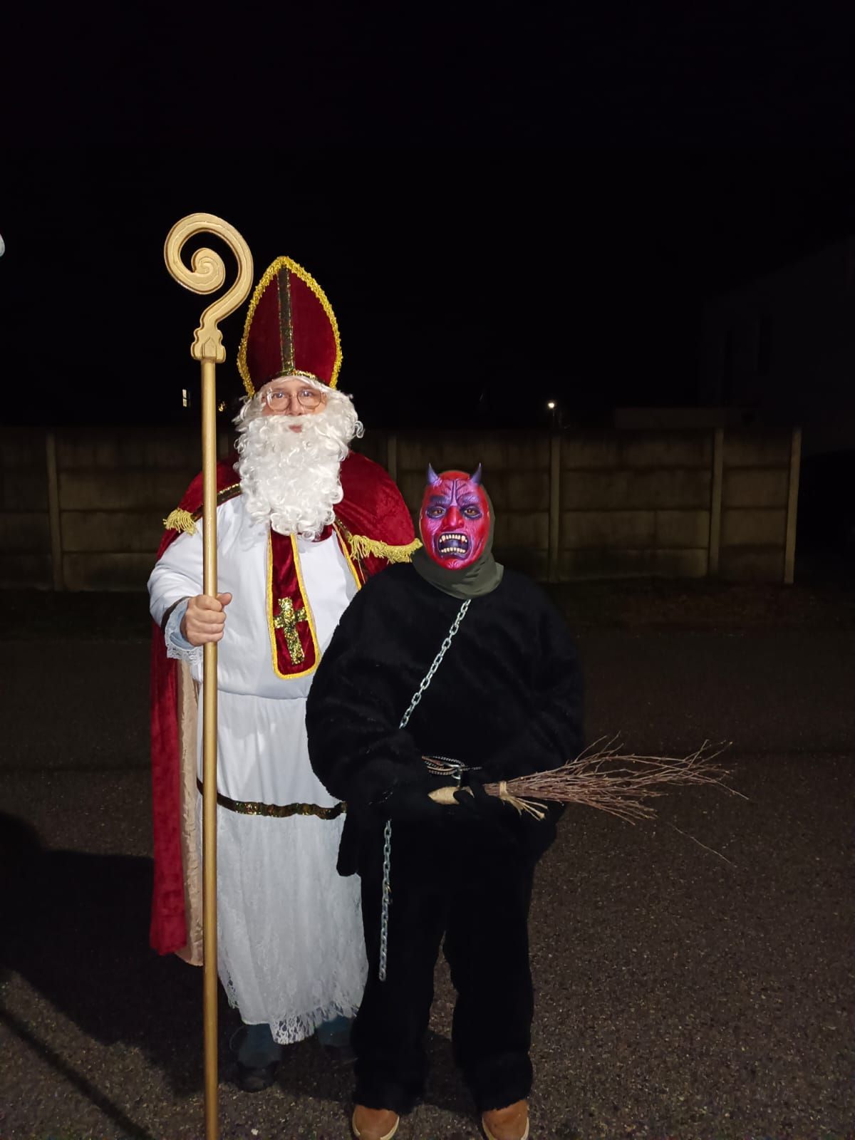 Ein Mann in der Kleidung des Heiligen Nikolaus steht neben einem anderen in Teufelskostüm, beide halten einen Besen, in einer nächtlichen Umgebung.