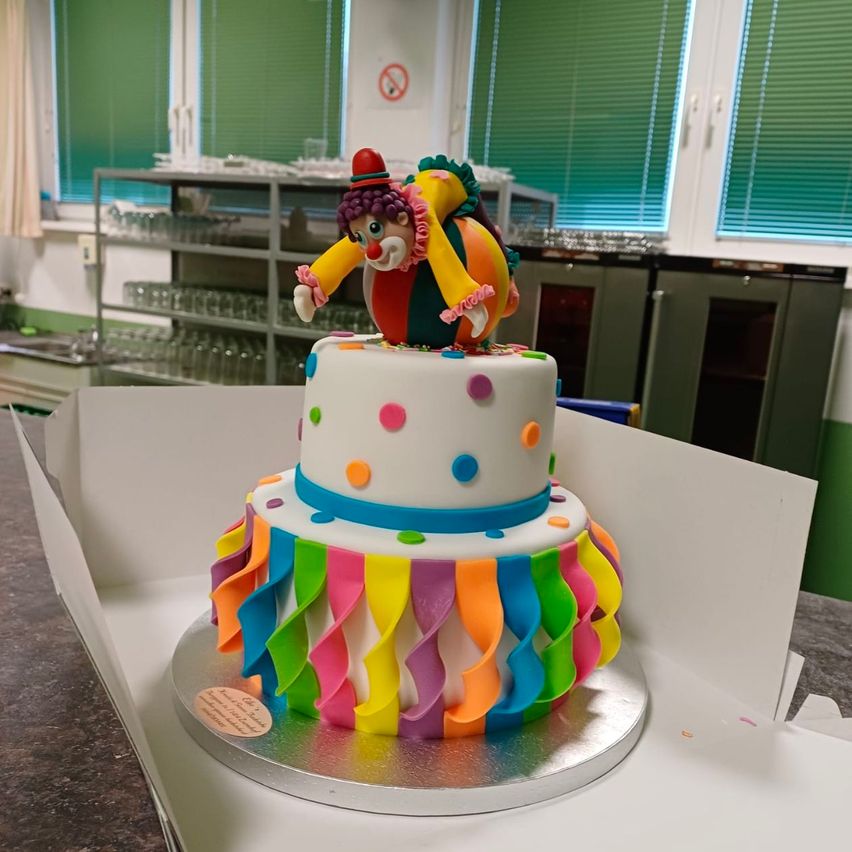 Ein farbenfroher Clown-Kuchen steht in einer Box auf einer Theke. Der Kuchen hat zwei Etagen, die jeweils mit bunten Bändern verziert sind. Obenauf steht eine Clown-Figur.