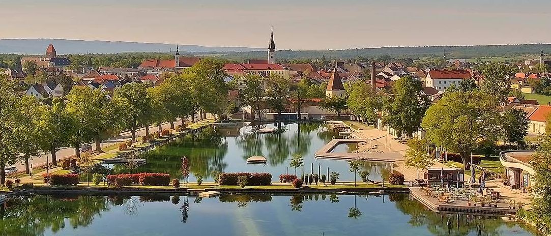 Luftaufnahme einer Stadt mit vielen Gebäuden, roten Dächern und einem großen See, umgeben von Bäumen und Grünflächen unter einem blauen Himmel mit Wolken.
