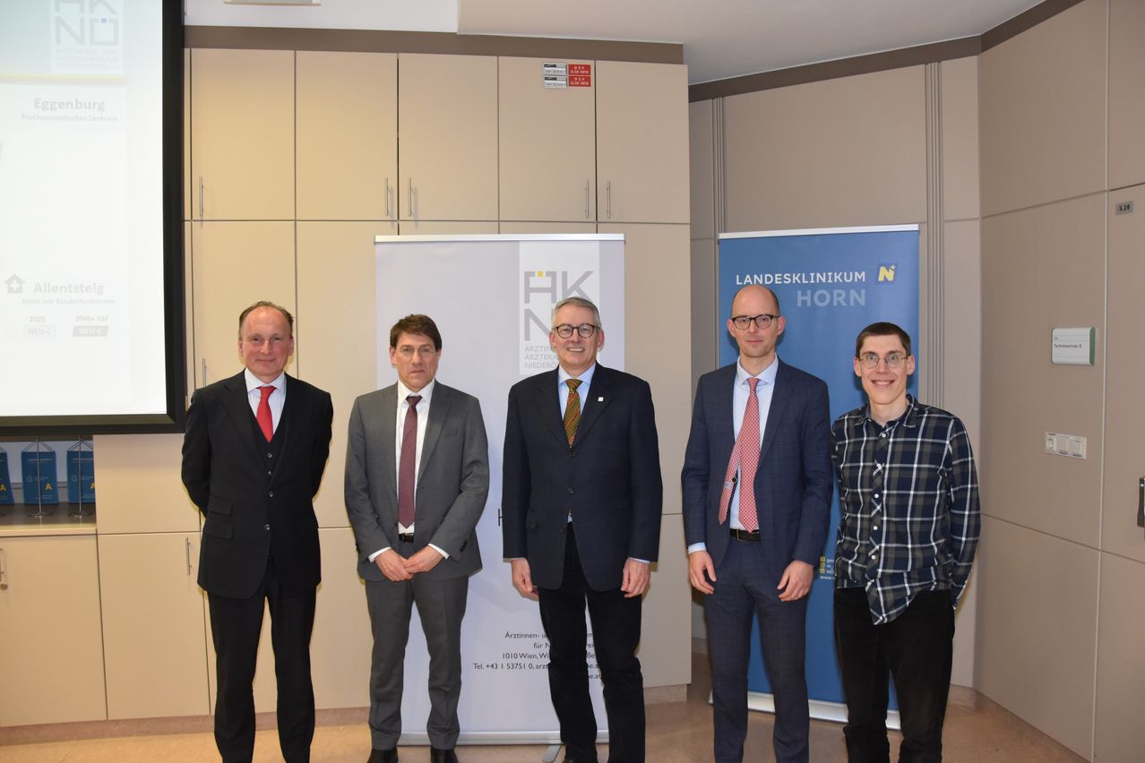 Fünf Männer in Anzügen und Krawatten stehen vor einem weißen Banner mit Text und einem blauen Banner.