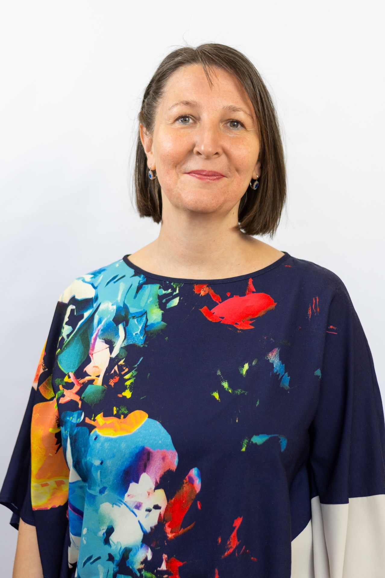 Porträt einer Frau, die ein marineblaues Hemd mit einem farbenfrohen abstrakten Design auf der Vorderseite trägt. Sie hat kurze, wellige braune Haare und lächelt in die Kamera. Der Hintergrund ist weiß.