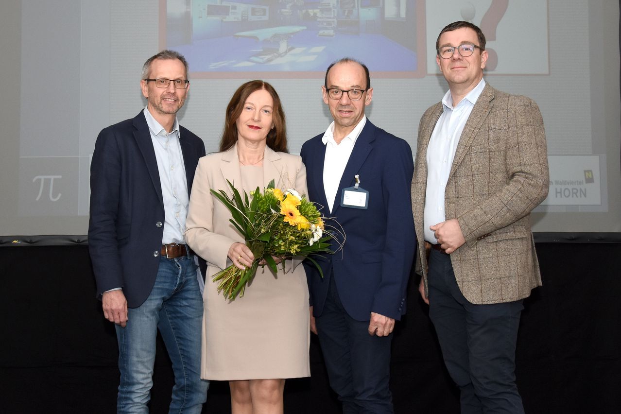 Vier Personen stehen eng beieinander, lächeln und schauen in die Kamera. Die Frau in der Mitte hält einen Blumenstrauß. Der Mann rechts trägt einen braunen Mantel, während die anderen beiden Männer blaue Anzüge tragen. Hinter ihnen befindet sich eine Projektionsleinwand mit einem Bild eines Krankenhauszimmers.