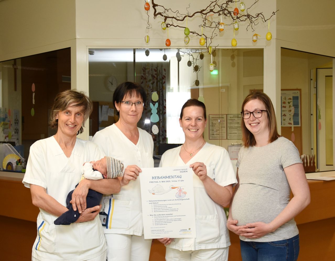 Vier Frauen stehen in einem Krankenhaus. Eine Frau hält ein Neugeborenes, und eine andere ist schwanger. Sie halten alle ein Plakat mit dem Text 'Hebammentag Freitag, 8. Mai 2015.' An der Wand hinter ihnen hängt eine Uhr.